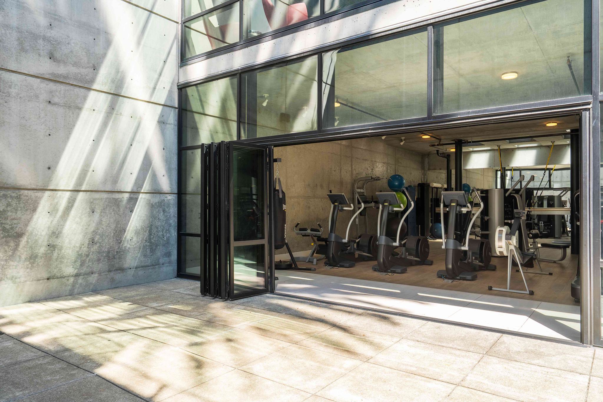 Indoor-outdoor fitness area at Alchemy with cardio machines and weight equipment, opening onto a private outdoor terrace through folding glass doors.
