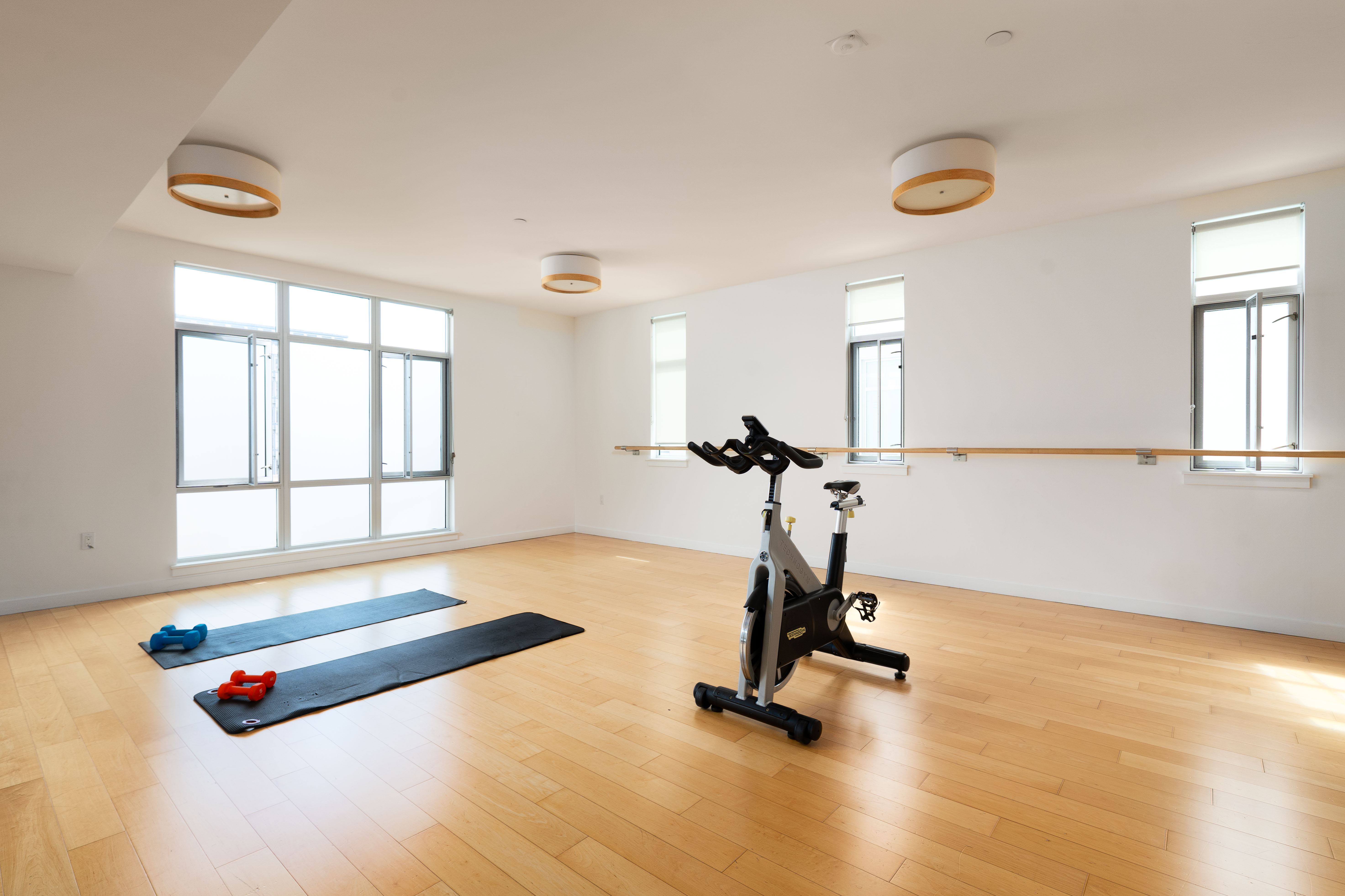 Bright spin and yoga studio at Alchemy with wood floors, large windows, a wall-mounted barre, exercise mats with weights, and a stationary bike.
