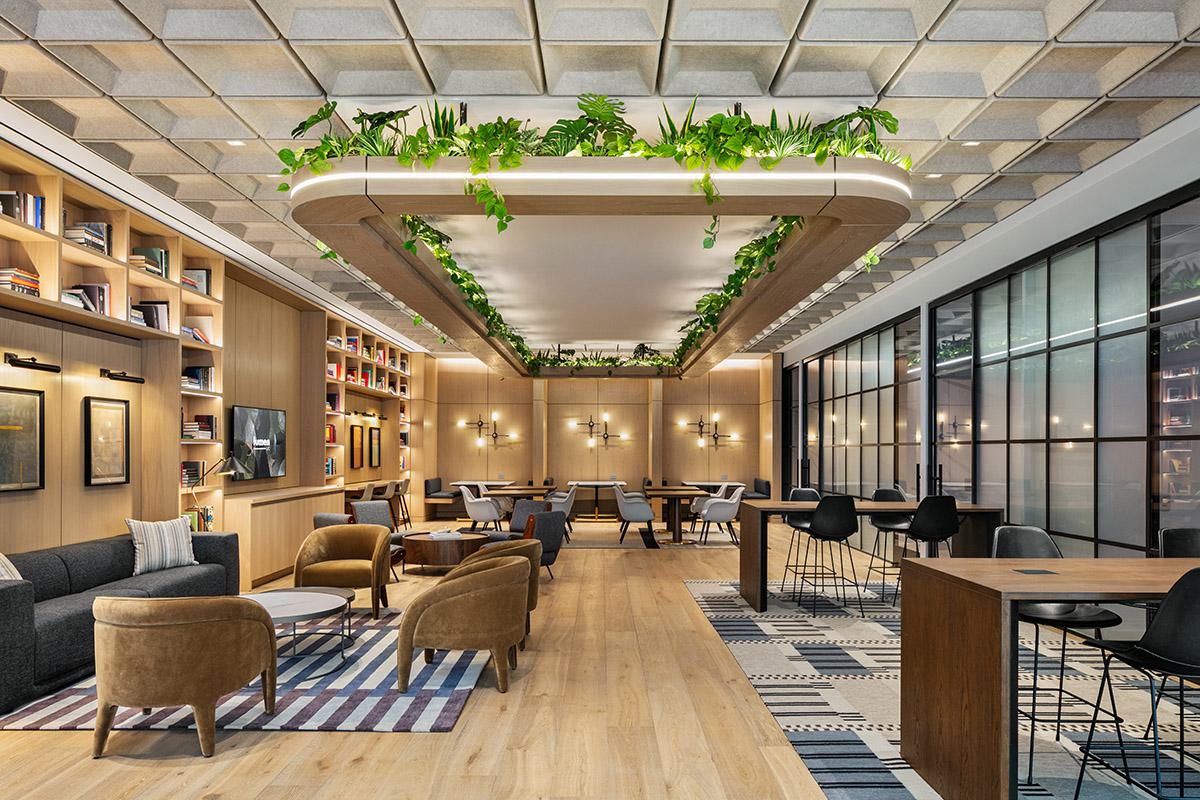 Coworking lounge with oak-paneled walls, built-in bookshelves, and a mix of seating. In the foreground, caramel-colored lounge chairs and a dark gray sofa sit around low round tables on striped rugs. Toward the back, communal tables and banquette seating are arranged under a ceiling installation edged with greenery. Along the right side, large black-framed glass panels enclose private conference rooms.