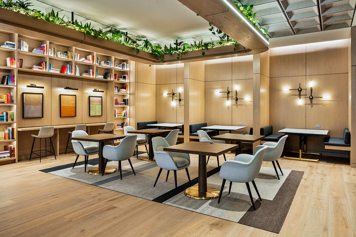 Coworking space with light wood floors and walls, lined with bookshelves and framed artwork. Several square tables with upholstered gray chairs are centered on area rugs, while booth-style banquette seating is built into the back wall. Overhead, a ceiling soffit trimmed with cascading plants adds greenery to the otherwise geometric design. Warm sconces provide soft, even lighting across the workspace.