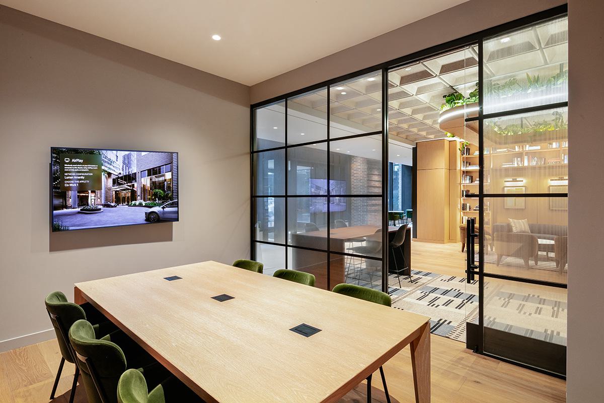 Private conference room at Lumen with a large wood table, green upholstered chairs, and a wall-mounted screen. Black-framed glass walls enclose the space, offering views into the main coworking lounge with bookshelves, seating, and greenery overhead.