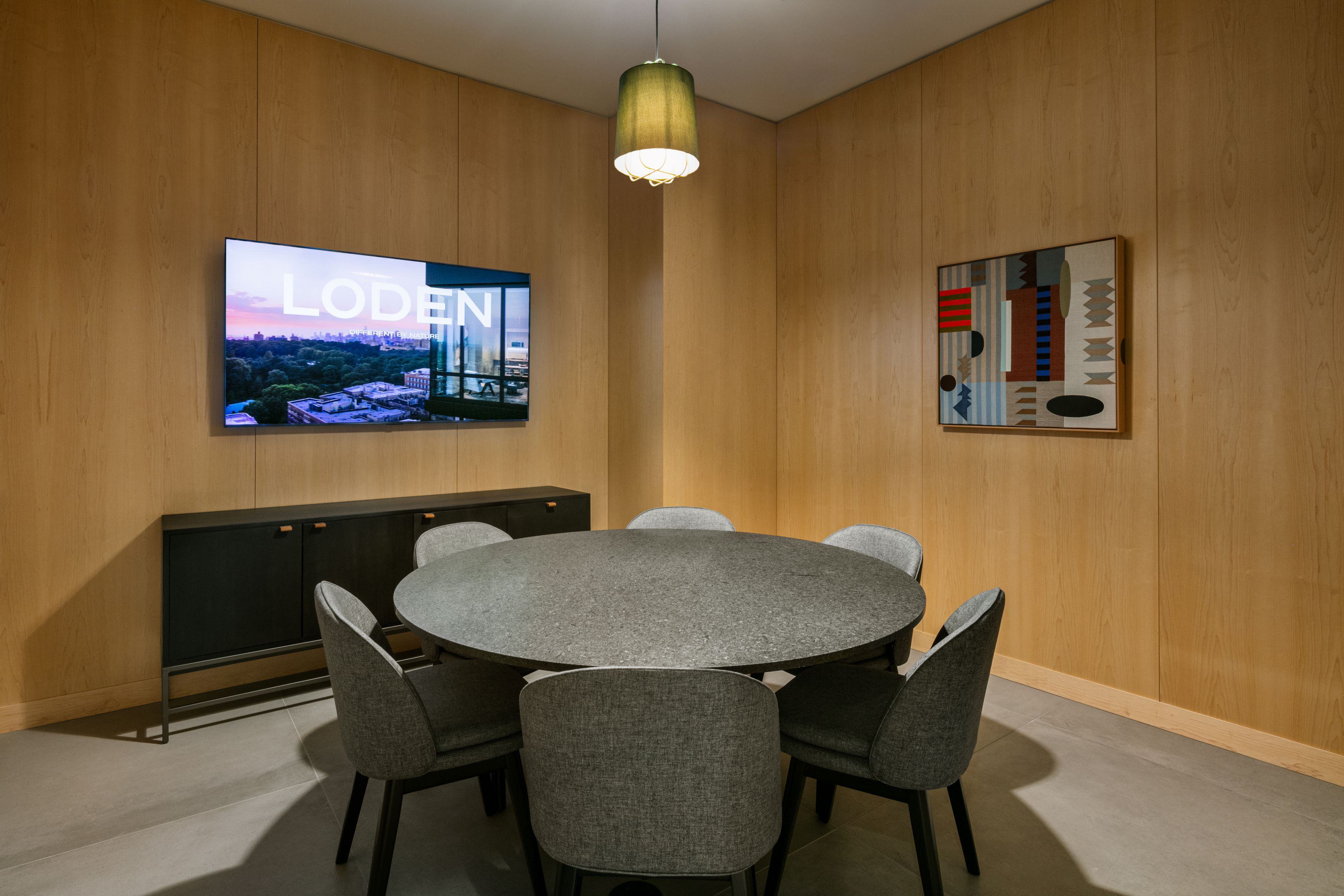 Private conference room at Loden with wood-paneled walls, a round stone table, upholstered gray chairs, and a wall-mounted screen display.