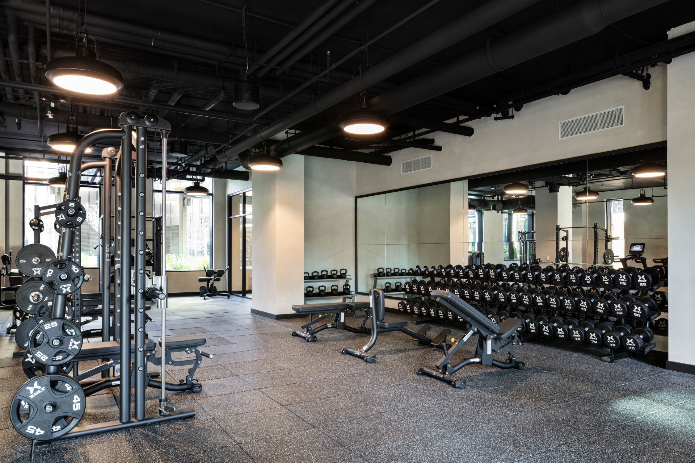 Strength training area at Loden featuring dumbbell racks, adjustable benches, squat rack, and cable machine against mirrored wall.