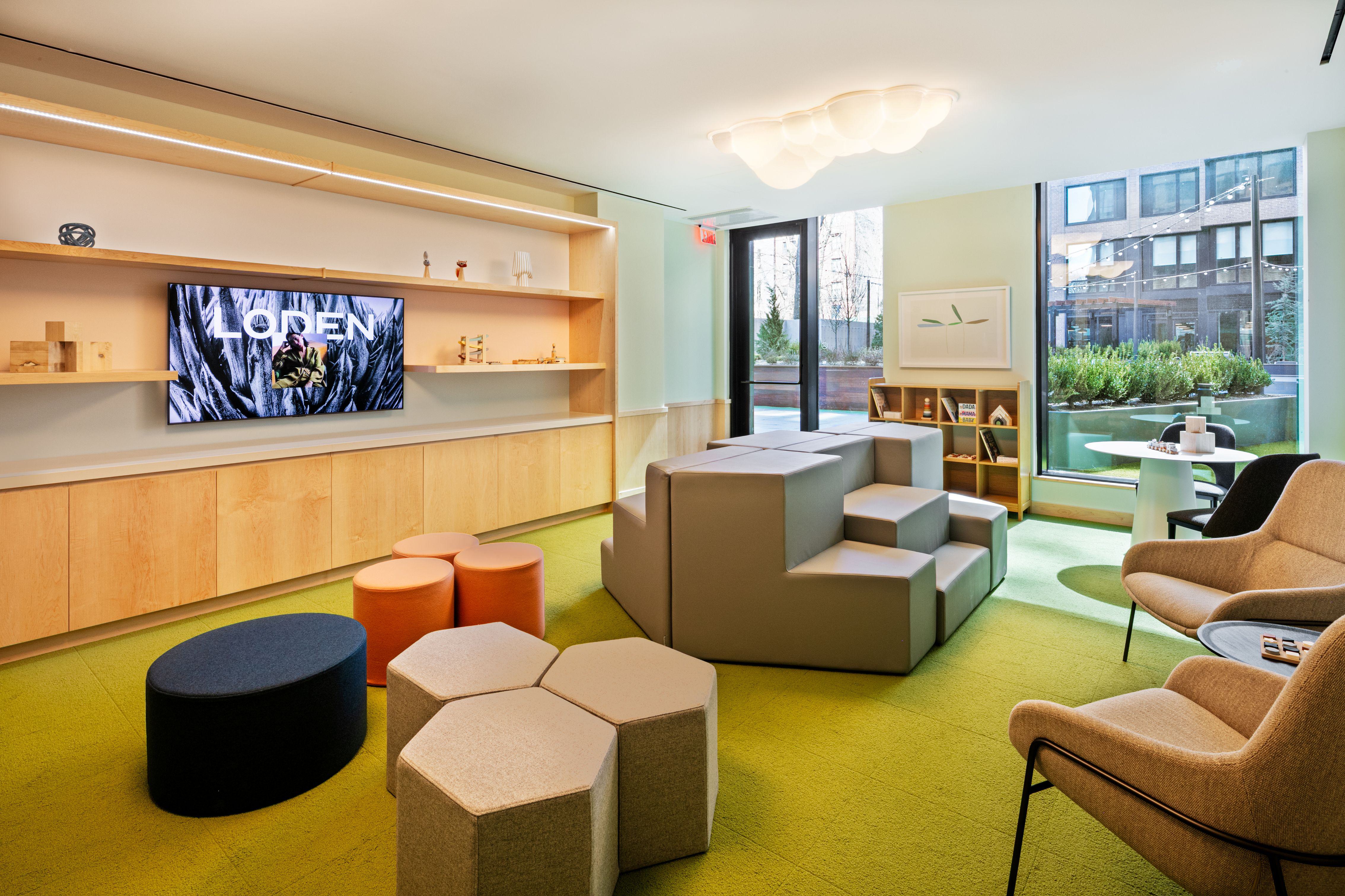Children’s playroom at Loden with colorful poufs, modular climbing blocks, modern chairs, bookshelves, and a wall-mounted TV facing green carpeted floors.