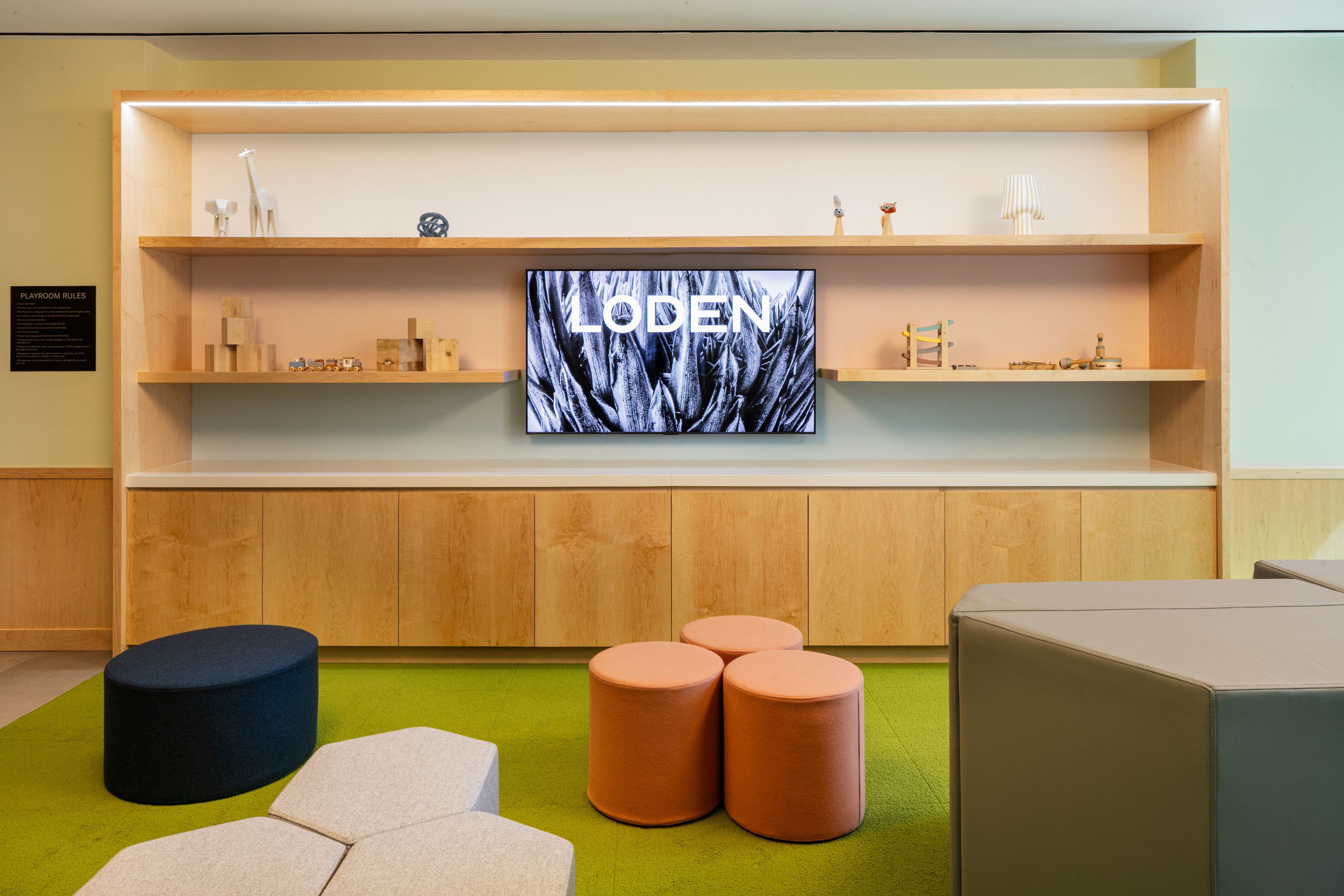 Playroom at Loden featuring a wall-mounted TV with wooden shelving, playful stools, and soft modular seating on a bright green floor.