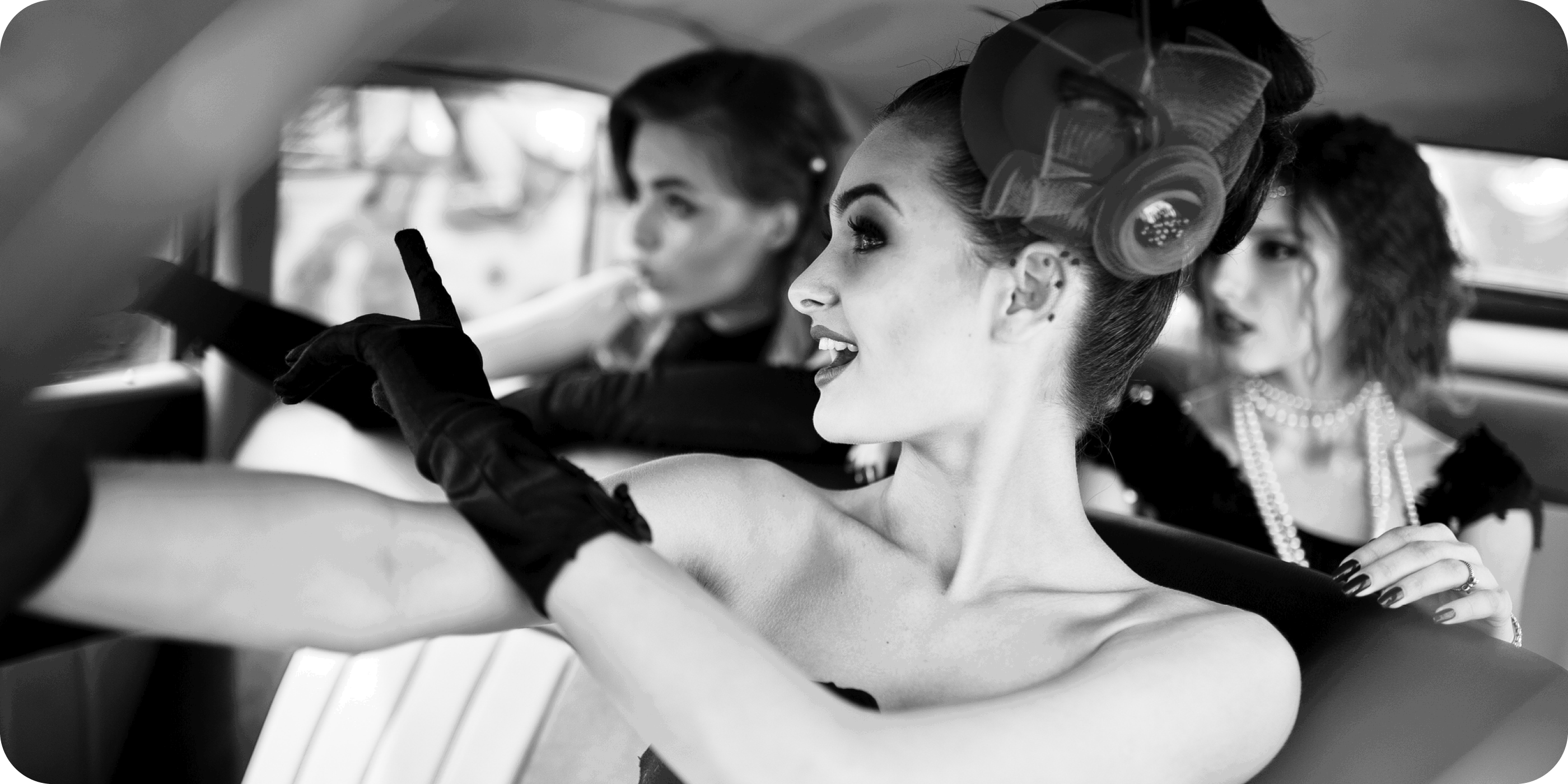 Black-and-white photo of elegantly dressed women in a vintage car, evoking classic Miracle Mile glamour.