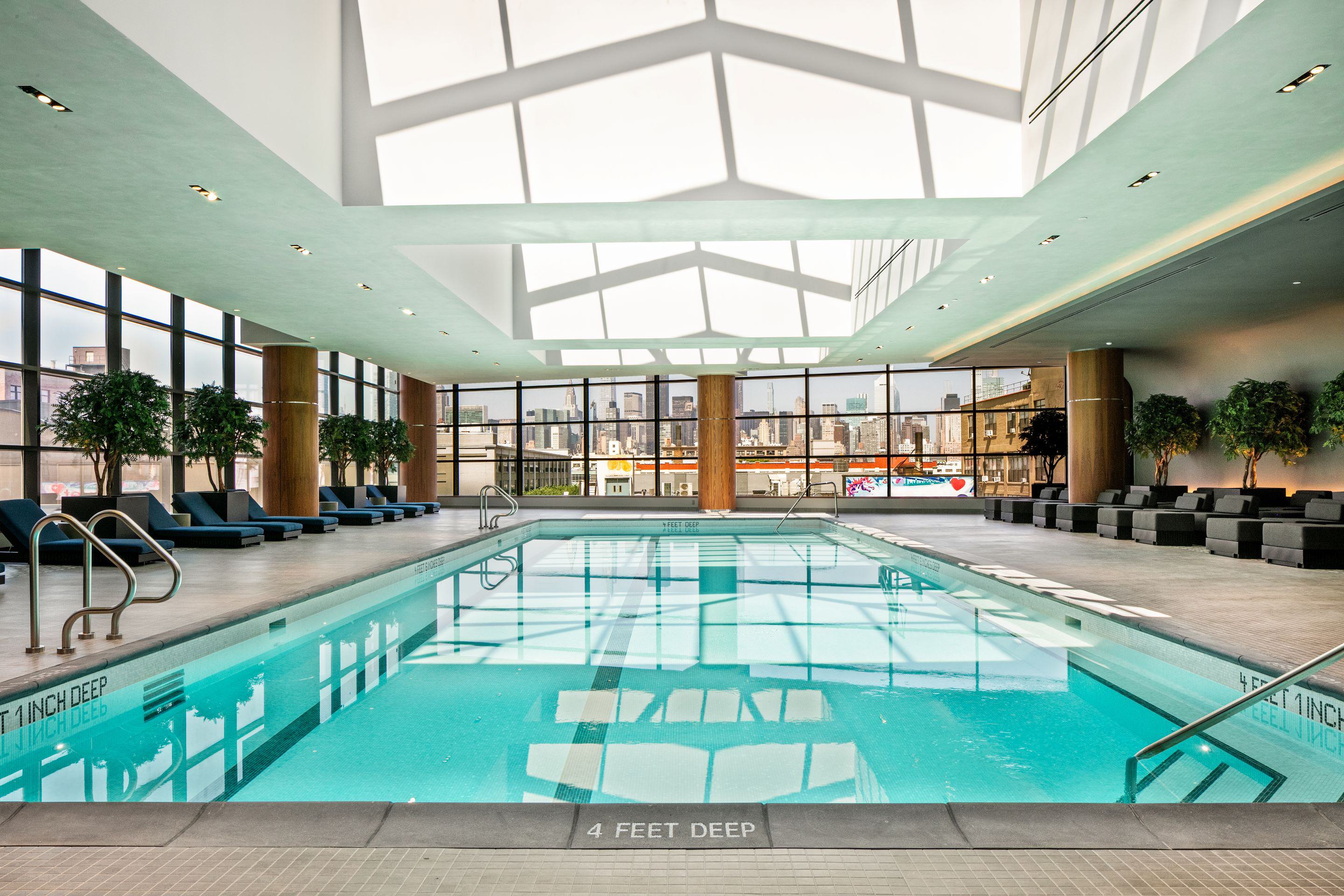 Indoor lap pool at Lumen LIC with skylit ceiling, floor-to-ceiling windows, and lounge seating overlooking the Manhattan skyline.