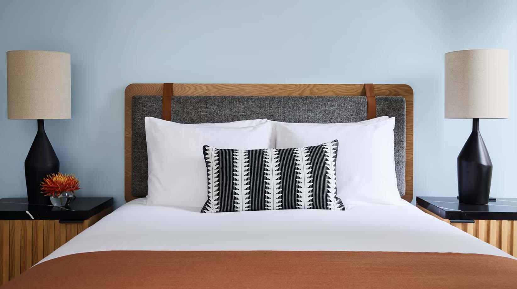 Modern furnished bedroom with a neatly made bed, geometric accent pillow, wood and fabric headboard, and matching black lamps on bedside tables.