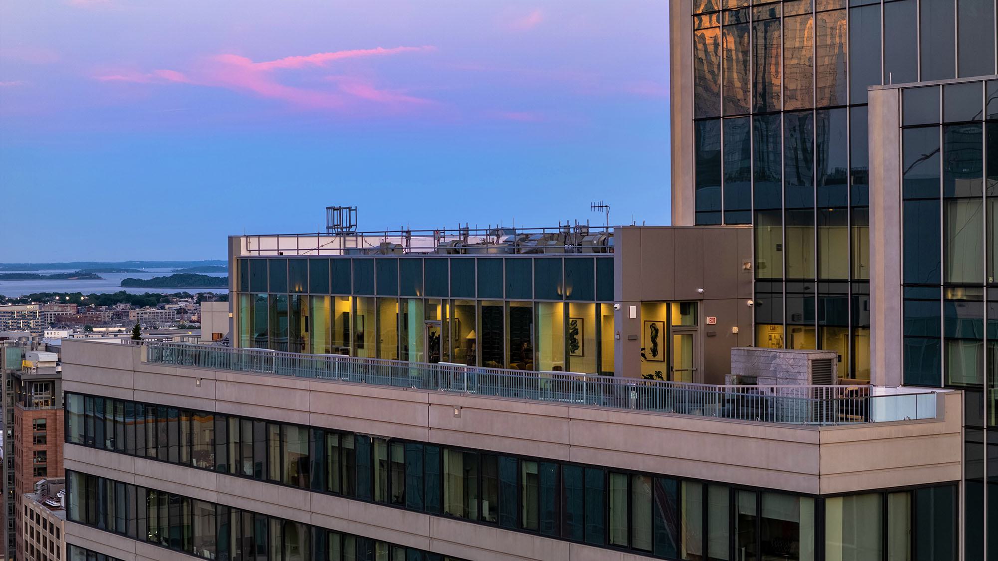 Exterior view of Luka on the Common’s rooftop lounge and terrace at sunset, showcasing glass walls, city reflections, and views toward Boston Harbor.