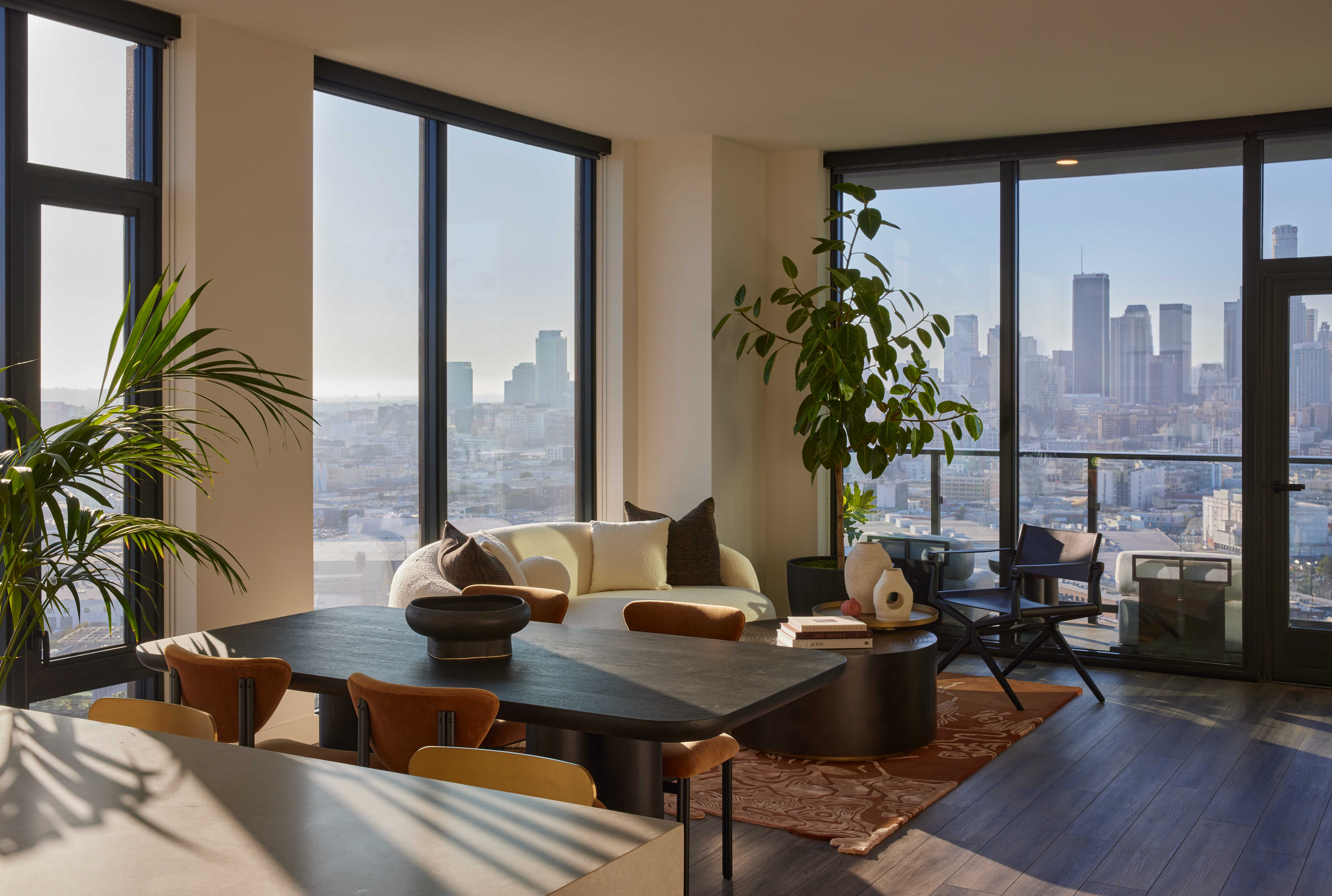 Modern high-rise apartment living room with floor-to-ceiling windows offering panoramic city views. The space features a curved cream sofa, dark wood dining table with tan leather chairs, large indoor plants, and warm natural light filtering through the expansive glass.