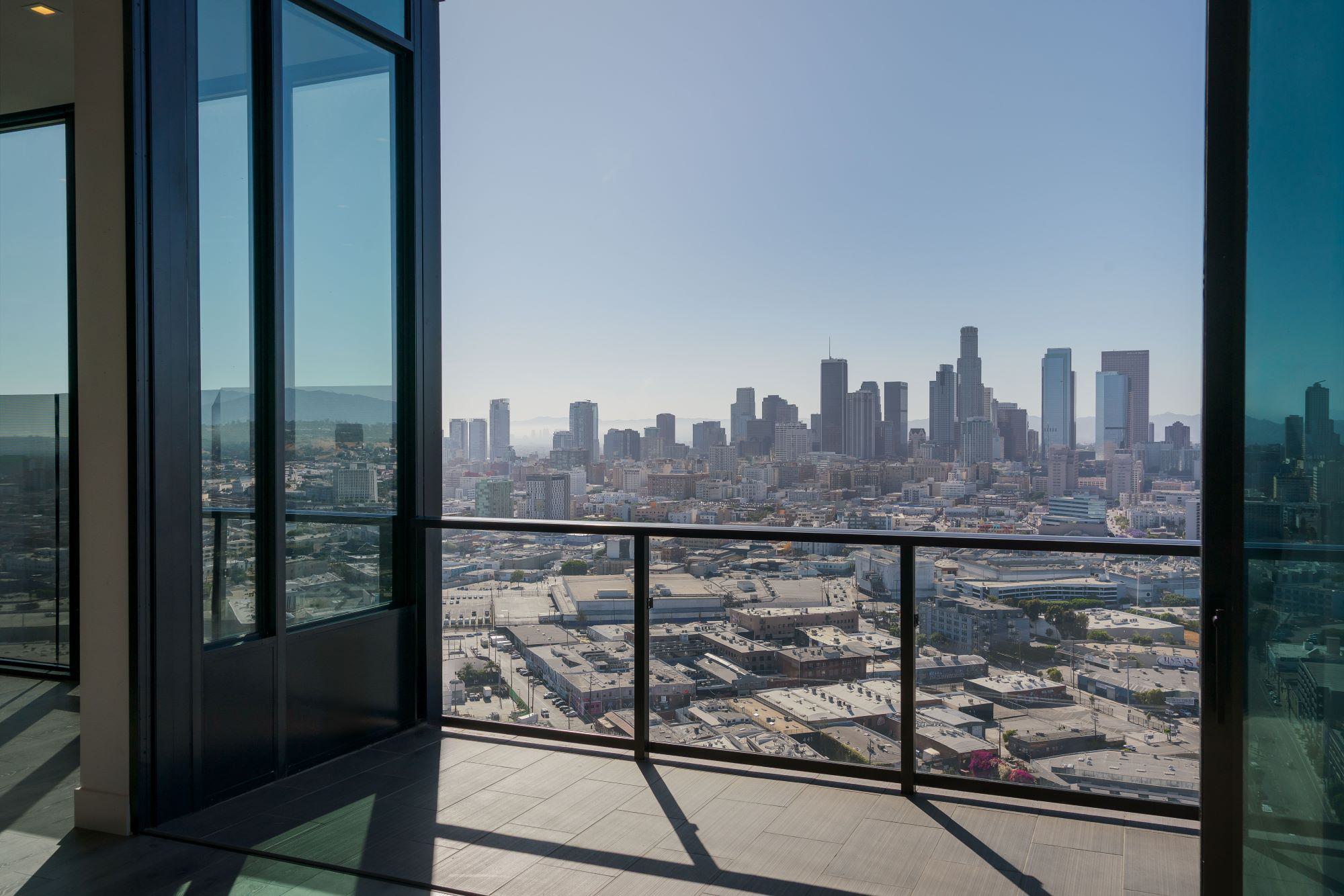 High-rise penthouse apartment balcony overlooking downtown Los Angeles. Expansive glass doors and railings frame a panoramic city skyline view under clear blue skies.