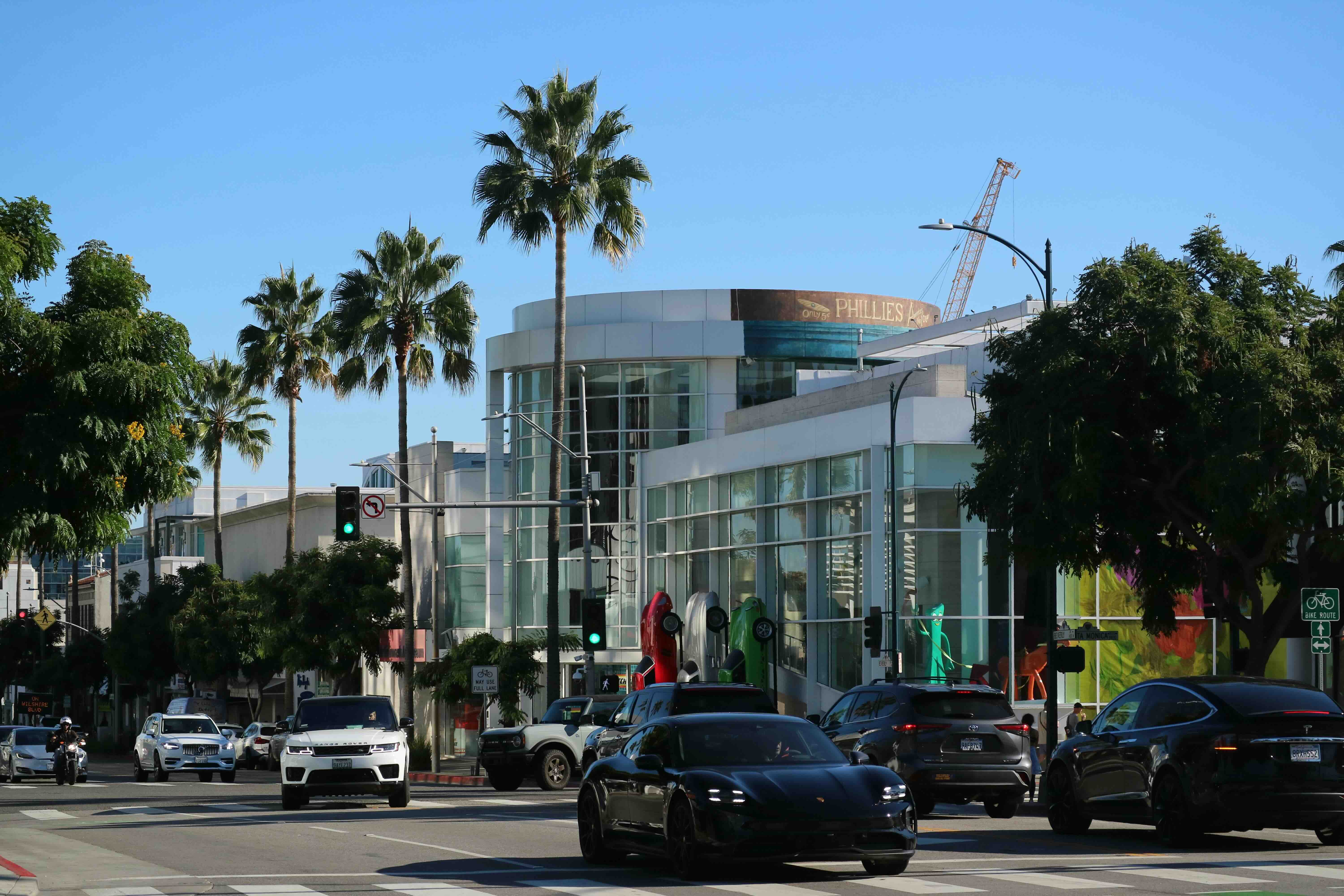 Street view of Wilshire Boulevard in Los Angeles’s Miracle Mile, showing cars at a busy intersection near the Petersen Automotive Museum. Modern glass buildings, palm trees, and colorful car sculptures line the street under a clear blue sky.