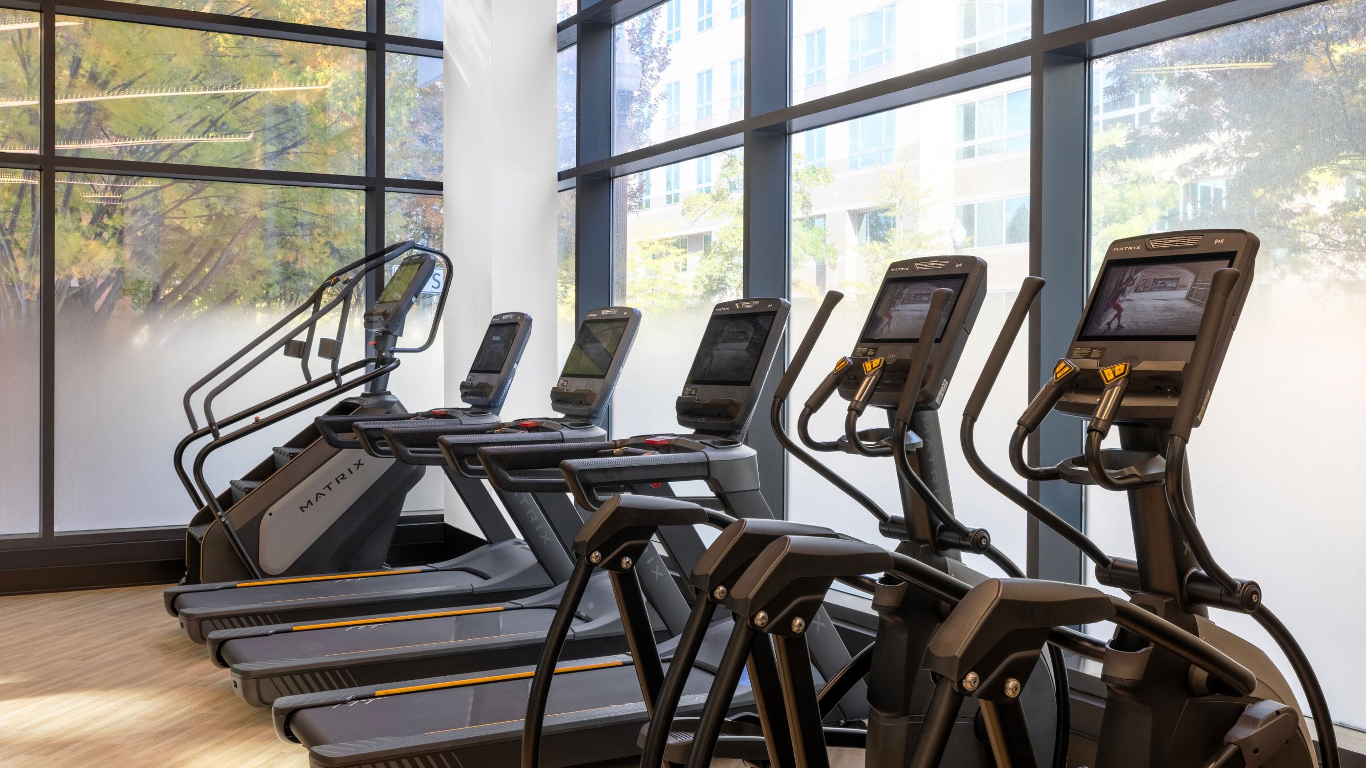Resident fitness center at Latitude Apartments in Virginia Square with modern cardio equipment, including treadmills and ellipticals, set along floor-to-ceiling windows with natural light and tree-lined views.