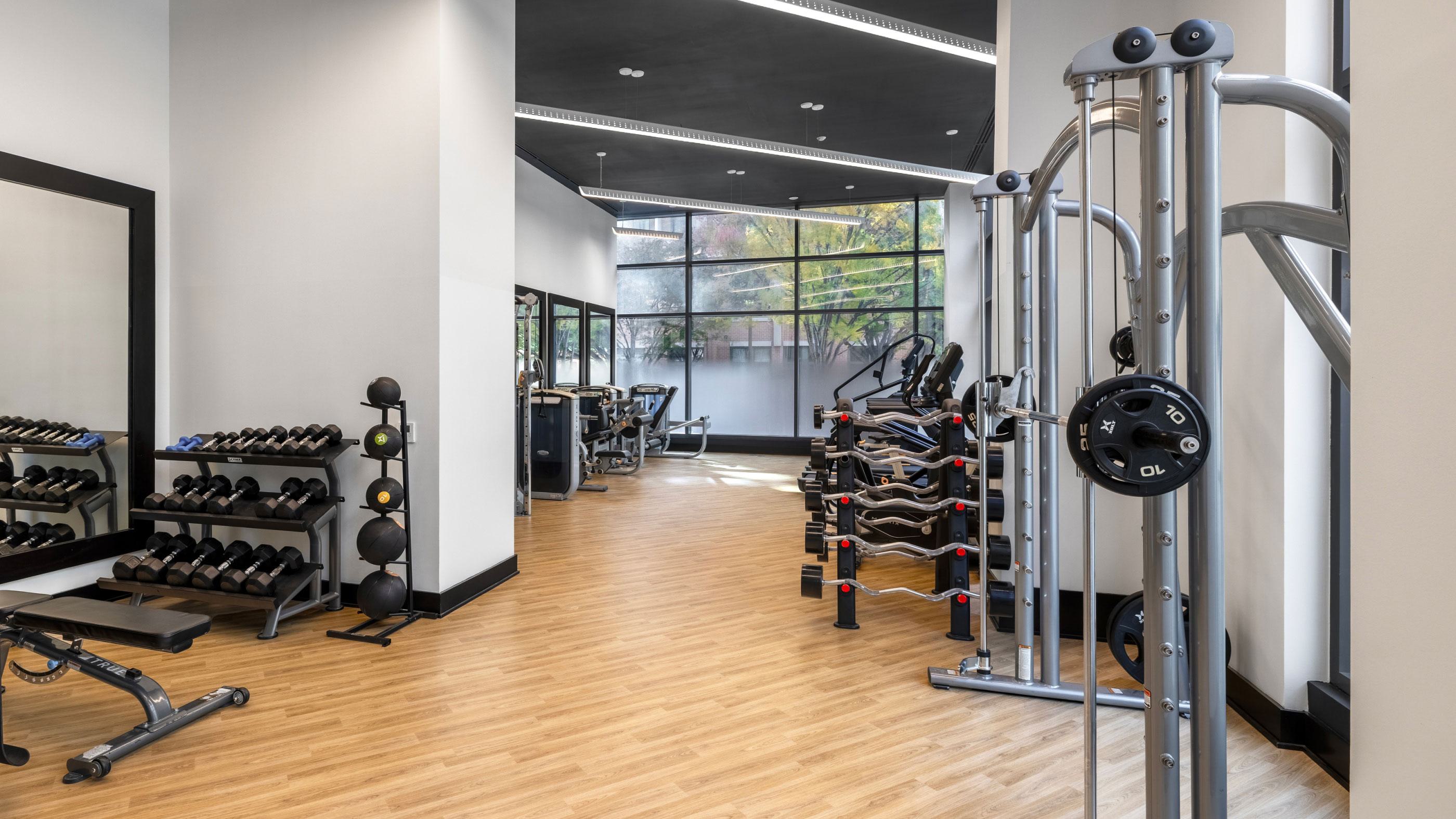 Resident fitness center at Latitude Apartments in Virginia Square with free weights, strength-training equipment, wood flooring, and floor-to-ceiling windows overlooking trees.