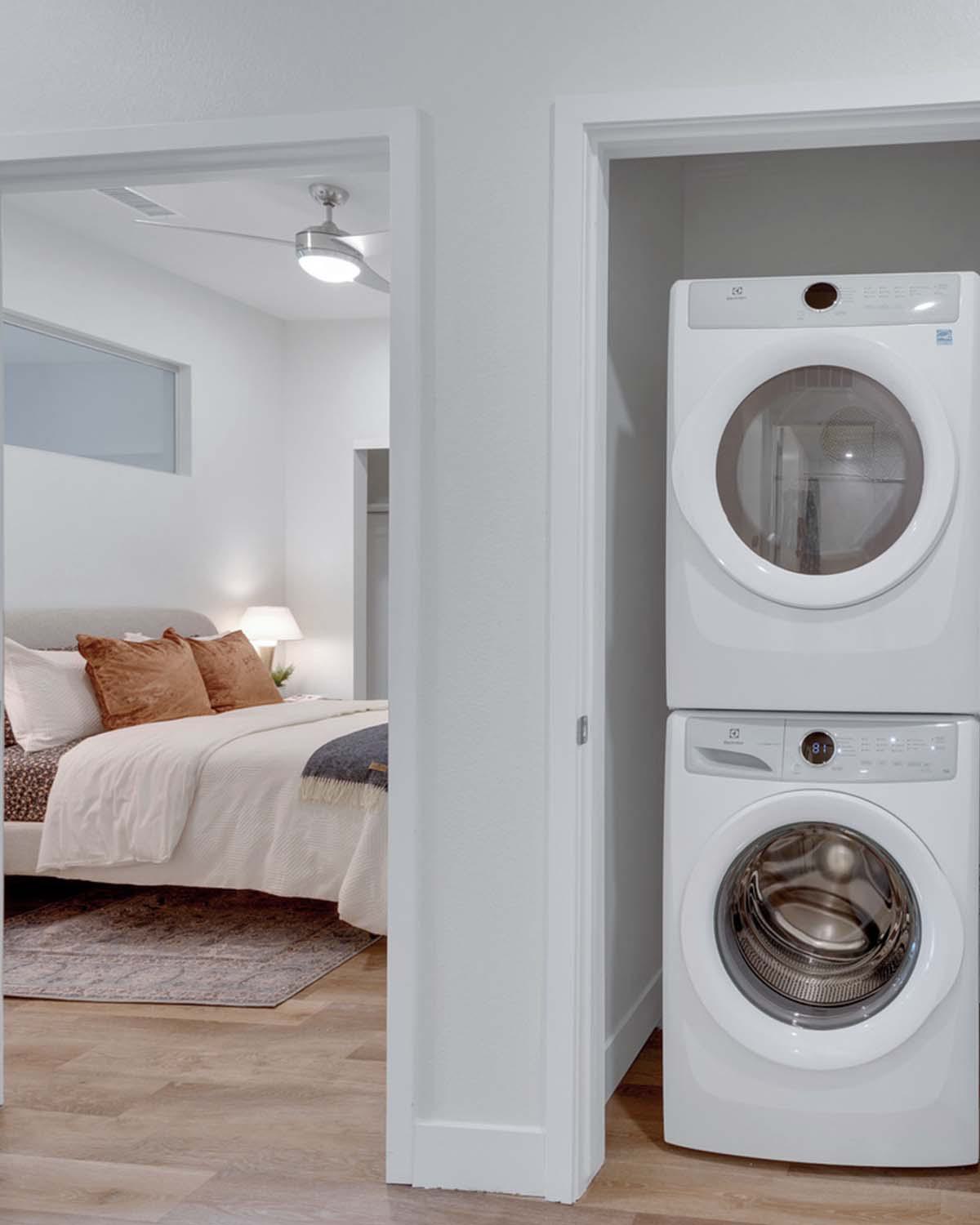 In-home stacked washer and dryer closet at Latitude Apartments in Arlington, VA, with a modern white laundry set, adjacent walk-in closet storage, and a glimpse into a bright, softly furnished bedroom beyond.
