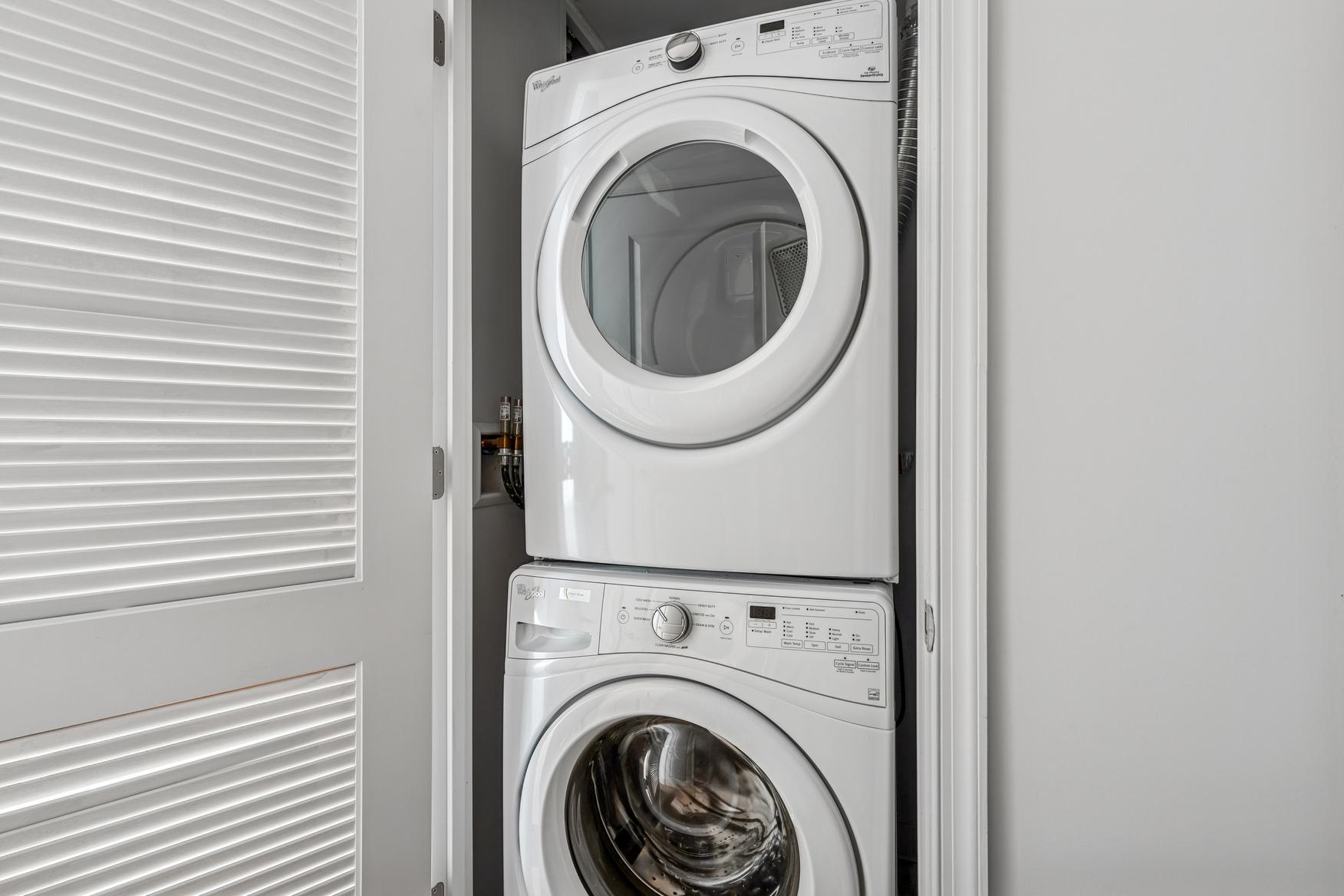 In-home stacked washer and dryer closet at Latitude Apartments in Arlington, VA, with a modern white laundry set, adjacent walk-in closet storage, and a glimpse into a bright, softly furnished bedroom beyond.