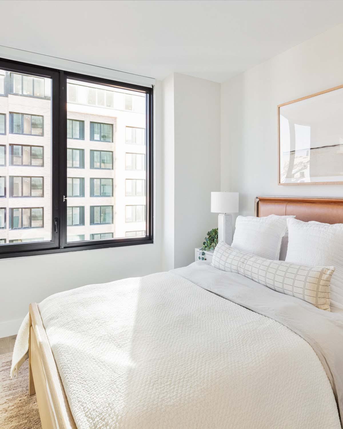 Bright, modern bedroom at Latitude Apartments in Arlington, VA, featuring a leather-paneled headboard, layered neutral bedding, bedside tables with contemporary lamps, and a large black-framed window overlooking neighboring buildings.
