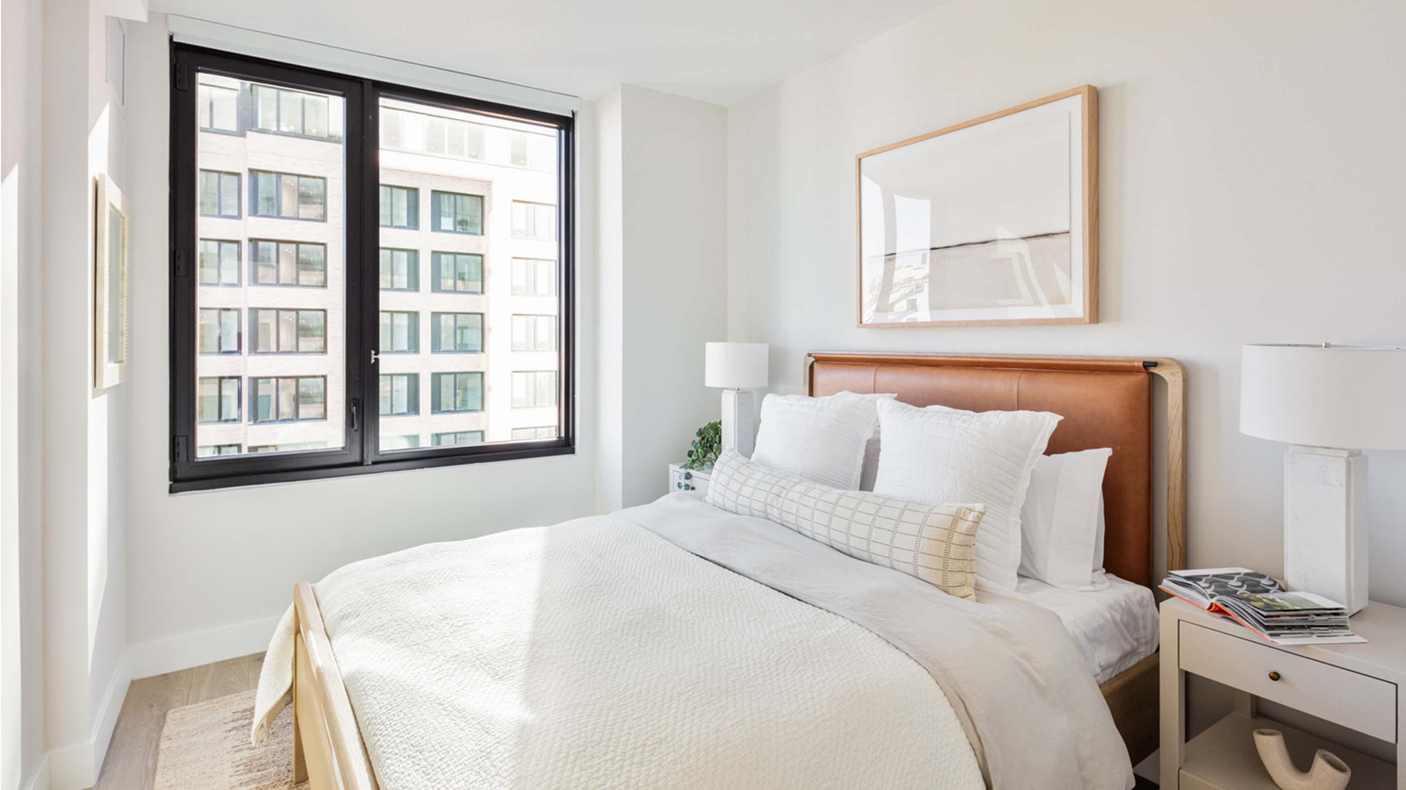 Bright, modern bedroom at Latitude Apartments in Arlington, VA, featuring a leather-paneled headboard, layered neutral bedding, bedside tables with contemporary lamps, and a large black-framed window overlooking neighboring buildings.