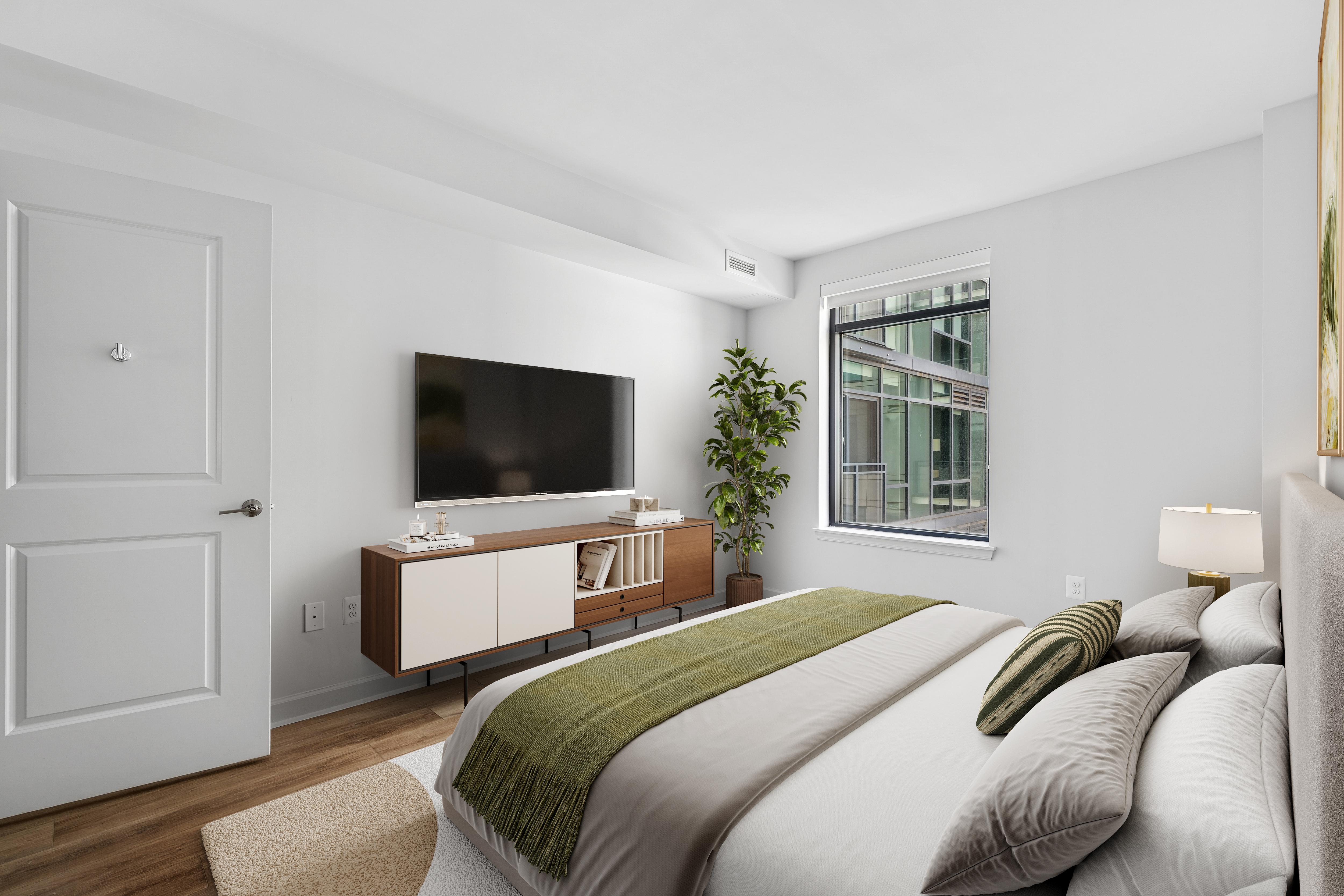 Bright, modern bedroom at Latitude Apartments in Arlington, VA, featuring a leather-paneled headboard, layered neutral bedding, bedside tables with contemporary lamps, and a large black-framed window overlooking neighboring buildings.
