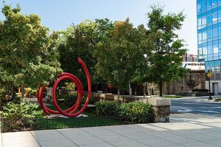 Curving red steel sculptures accent a small urban park in NoMa, Washington DC, surrounded by leafy trees and modern glass buildings, reflecting the neighborhood’s blend of art and contemporary design.