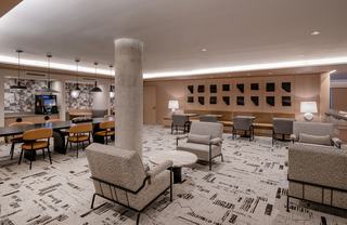 A sophisticated coworking lounge featuring neutral tones, modern furnishings, and layered textures. The space includes upholstered armchairs with metal frames, a long communal table with caramel leather chairs, pendant lighting, and geometric wall art. A coffee station and patterned carpeting add comfort and visual interest, creating a warm, contemporary work environment