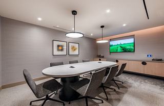 A private meeting room at The Hale featuring a light stone table, gray upholstered chairs, and a built-in wall display. Glass walls framed with vertical wood slats enclose the space, while a modern pendant light and neutral wall tones create a calm, refined atmosphere for meetings or focused work.