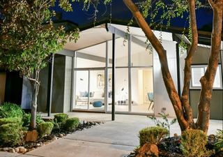 Exterior of a mid-century modern rental home at The Arbors in Antelope, California, featuring a sharply angled roofline, tall floor-to-ceiling windows, and warm interior lighting visible through the glass façade surrounded by minimalist landscaping and mature trees at dusk.
