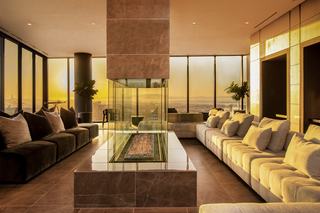 Sunset Lounge at Atlas Oakland featuring modern sofas, a glass fireplace, and panoramic floor-to-ceiling windows overlooking downtown and the Bay at sunset.