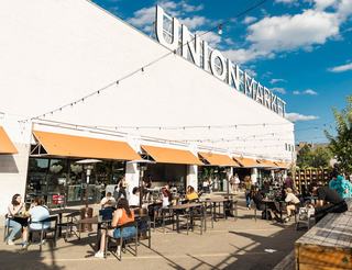 Outdoor dining area in front of the Union Market building in Washington, D.C., showing people seated at tables under orange awnings and string lights on a sunny day.