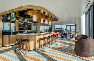 Indoor rooftop lounge bar area at Theory featuring a curved marble-topped bar with brass canopy lighting, modern barstools, colorful geometric tile flooring, and floor-to-ceiling windows overlooking the city and rooftop terrace.