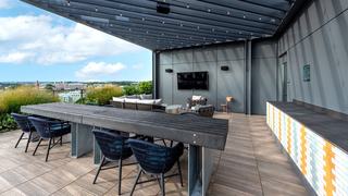 Outdoor dining and grill area at Theory featuring a modern pergola, long dining table with blue woven chairs, lounge seating, counter with sink, and city views.