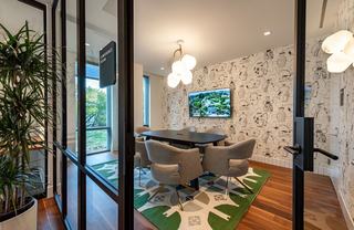 Glass-walled conference room at Theory with patterned wallpaper, green rug, gray upholstered chairs, oval table, sculptural pendant light, and wall-mounted screen.