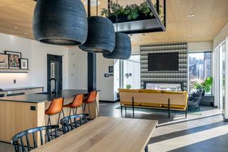Rooftop sky lounge at Arlo Apartments featuring a modern kitchen with wood cabinetry, black countertops, pendant lighting, a fireplace, TV, and floor-to-ceiling windows opening to the outdoor deck.