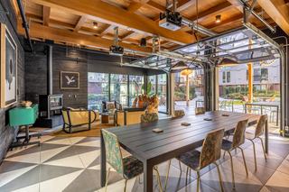 Modern indoor pavilion at Arlo Apartments featuring a long dining table, rattan lounge seating, geometric tile floors, exposed wood ceiling beams, fireplace, and glass garage-style doors that open to the courtyard patio.