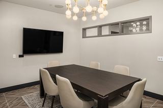 A modern conference or meeting room with a dark wood table and six cream-colored chairs, a large wall-mounted TV, and a contemporary brass chandelier with globe lights above. The neutral walls and minimal decor give the space a clean, professional look.