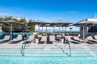 Rooftop pool deck at Linea featuring a rectangular pool surrounded by gray cushioned lounge chairs, large charcoal umbrellas, and blue tile accents, with panoramic views of Century City, the Hollywood Hills, and distant San Gabriel Mountains under a clear Los Angeles sky.