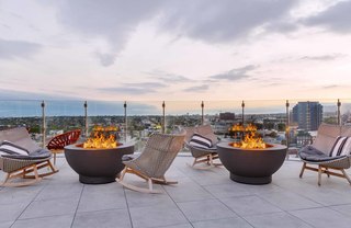 Rooftop fire pit lounge at Linea featuring round modern fire bowls surrounded by cushioned woven chairs with striped pillows, overlooking the Los Angeles skyline at sunset through glass railings.