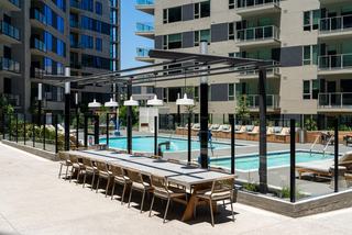 Open-air courtyard at Linea Apartments with a long outdoor dining table under a modern pergola, overlooking a pool enclosed by glass railings and lined with built-in wood daybeds and lounge chairs.