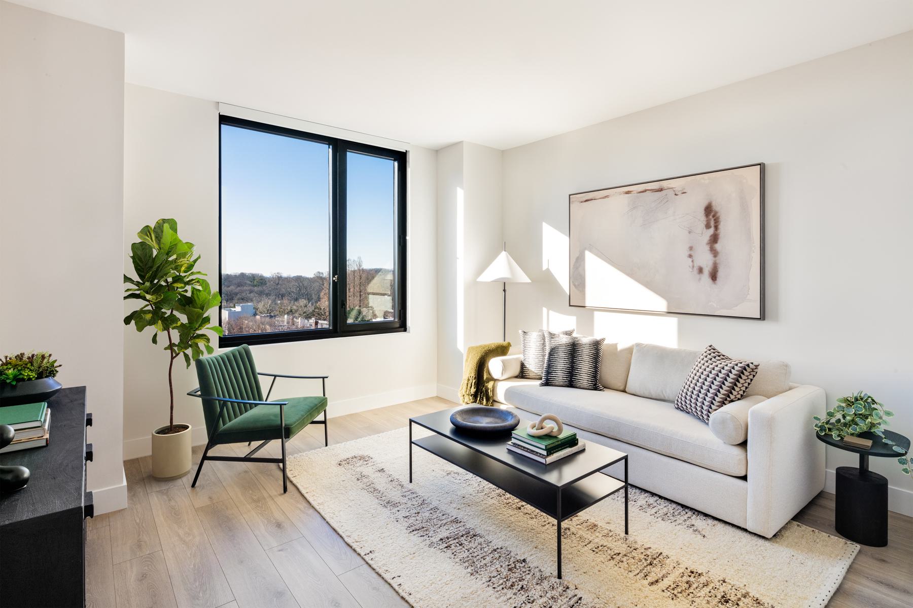Sunlit living room at Loden with large black-framed windows, a cream sofa, green accent chair, and views of treetops and surrounding Brooklyn buildings.