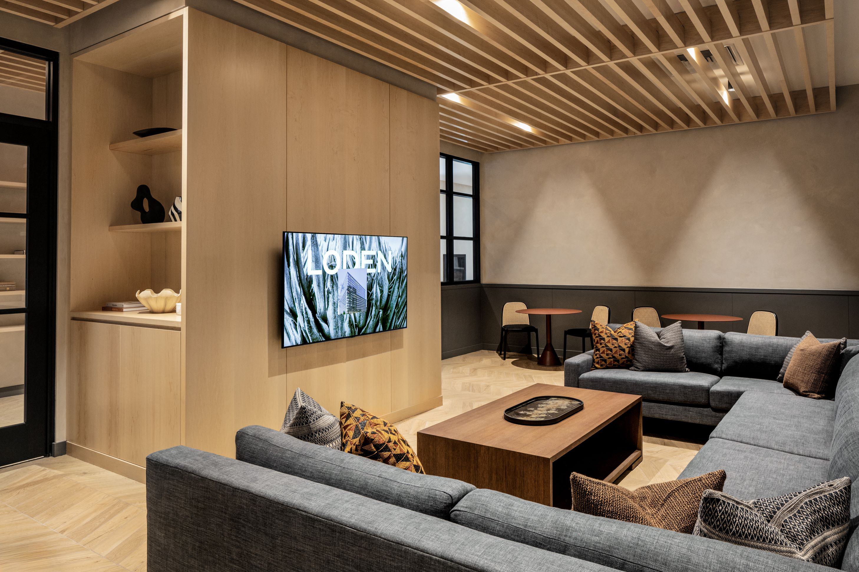 Loden TV lounge with wraparound seating, accent pillows, built-in shelving, wood ceiling slats, and café-style tables along the back wall.