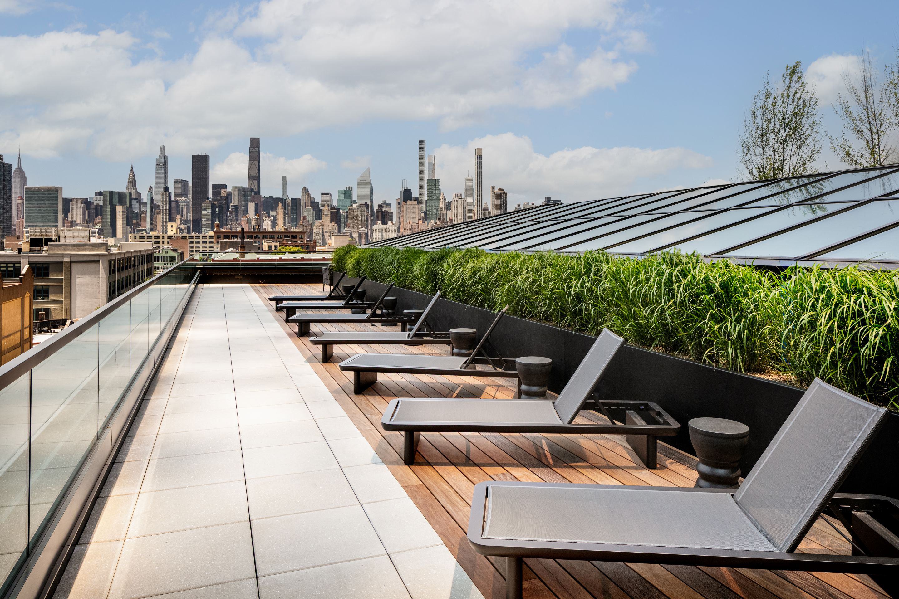Rooftop sun deck at Lumen LIC featuring modern loungers, landscaped planters, glass railing, and unobstructed views of the Manhattan skyline.