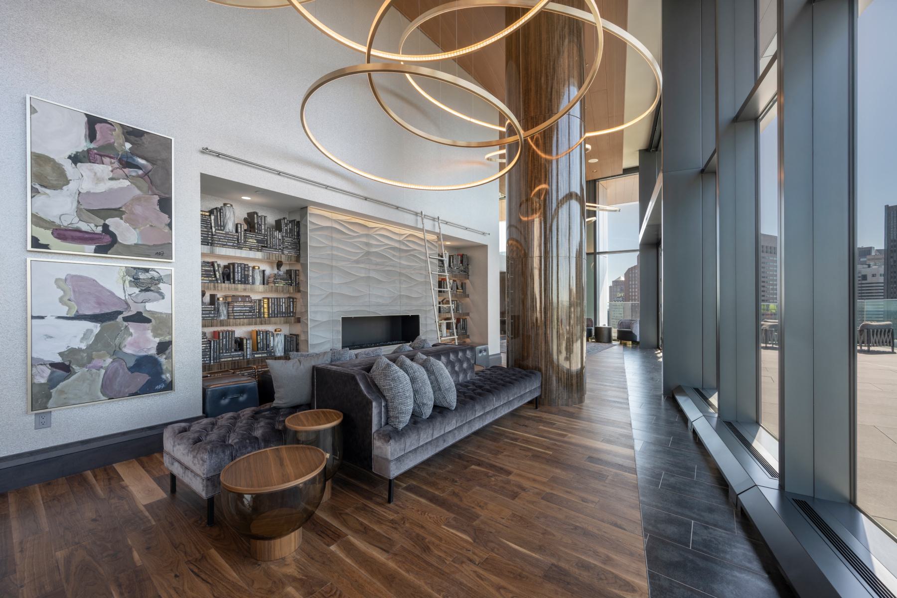Modern resident lounge at The Sudbury featuring floor-to-ceiling windows, sculptural ring lighting, a tufted velvet sofa, built-in bookshelves with a rolling library ladder, and a textured fireplace wall.