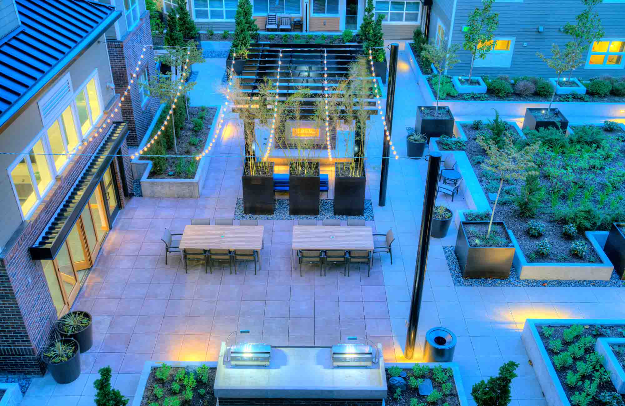 Aerial view of Urbana’s courtyard at dusk, with glowing string lights, grill stations, long dining tables, modern planters, and lush landscaping arranged around a central fireplace pavilion.