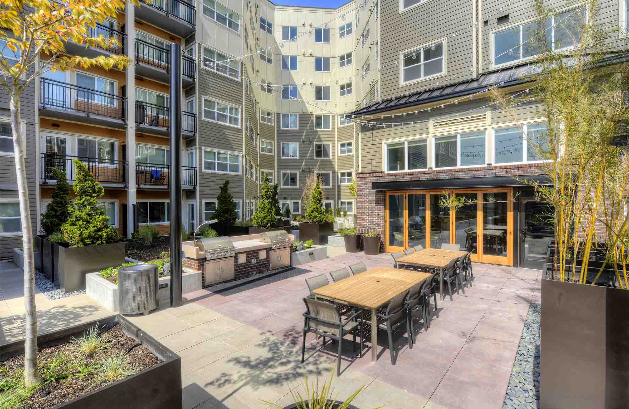 Daytime view of Urbana’s courtyard featuring communal dining tables, stainless-steel grills, tall planters with greenery, and surrounding apartment balconies.