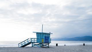 A pastel blue lifeguard tower sits on a quiet stretch of beach at sunrise, with gentle waves, soft clouds, and distant coastal hills creating a calm Westside shoreline scene.