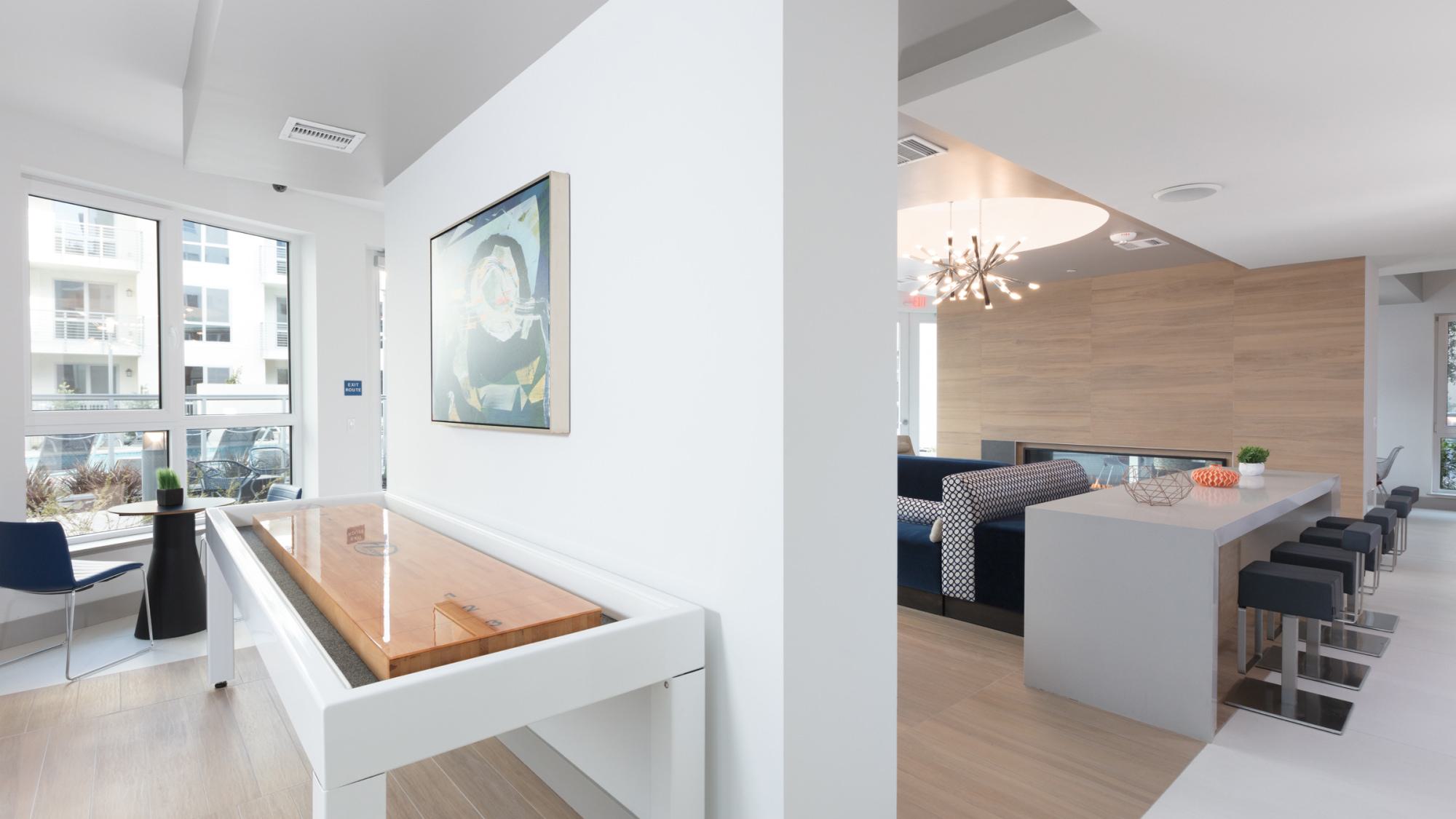 Bright clubhouse space at The Triton featuring a shuffleboard table, bar-height island seating, large windows, and adjacent lounge area.