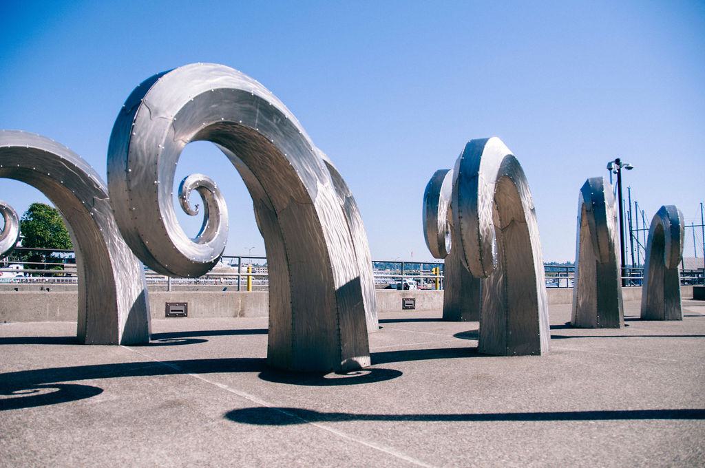 Stainless-steel “Salmon Waves” sculpture by Paul Sorey, featuring large curved metal forms arranged outdoors near the Ballard Locks against a clear blue sky.