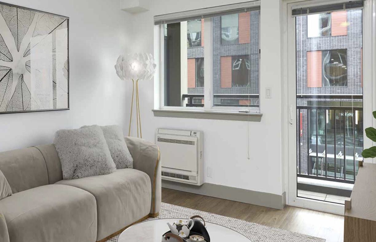 Bright living room at Waymark Apartments featuring a cozy sectional, woven media console, indoor plant, and sliding balcony door with urban views.