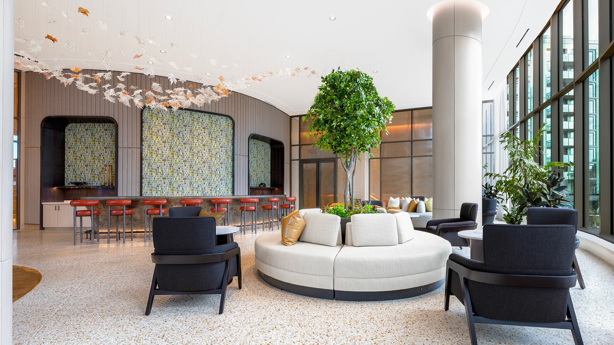 Aviary Lounge at Aerie featuring a circular cream sofa built around a large indoor tree, a curved bar with red stools and mosaic tile backsplash, tall columns, and full-height windows facing the courtyard.