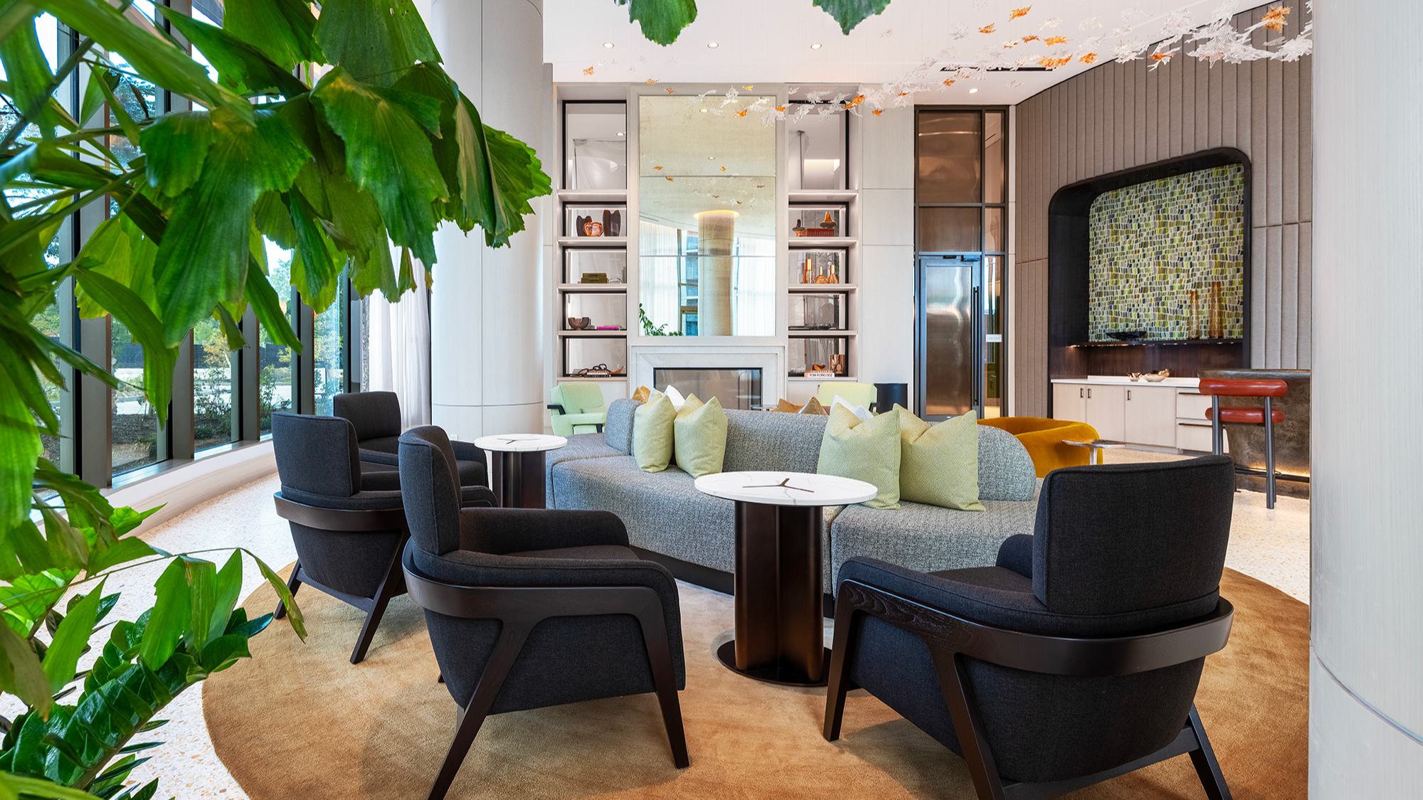 Aviary Lounge seating area at Aerie with dark lounge chairs, a long gray sofa with green pillows, a fireplace surrounded by open shelving, and floor-to-ceiling windows overlooking the courtyard gardens.